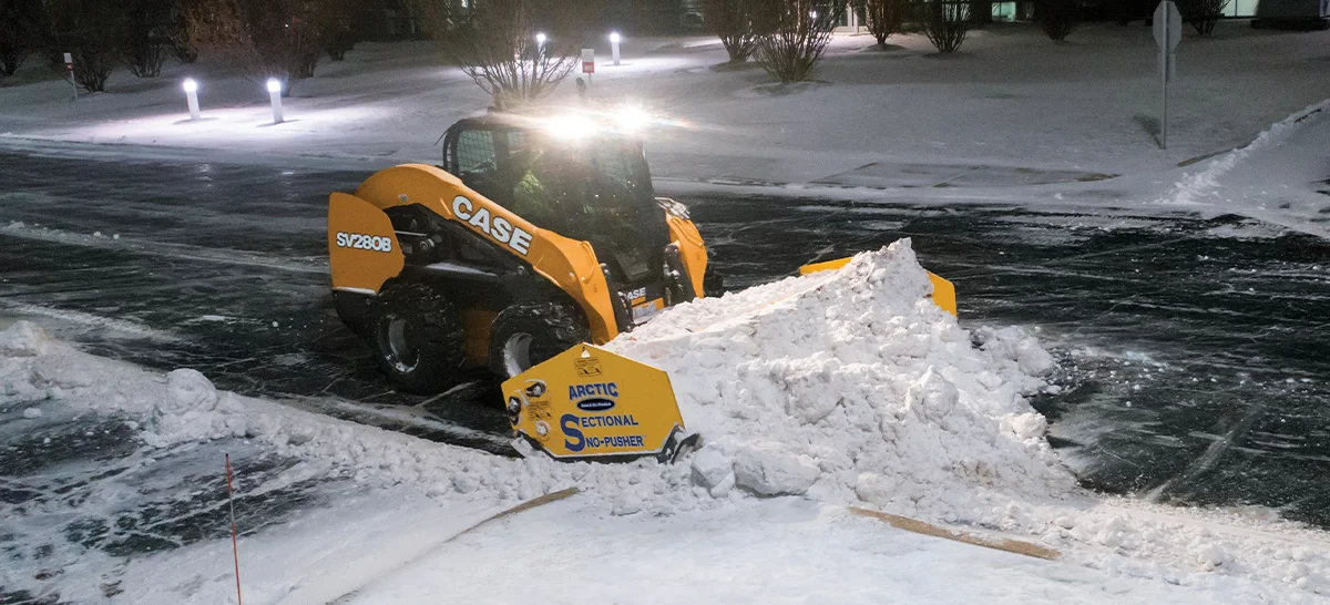 Case SV280B Skid Steer with a snow pusher attachment pushing snow on a street at night.